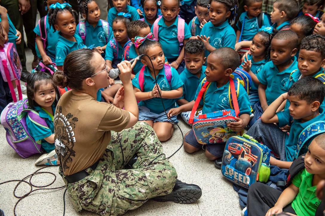 A sailor holding a microphone sits cross-legged on a floor with children and sings to them.