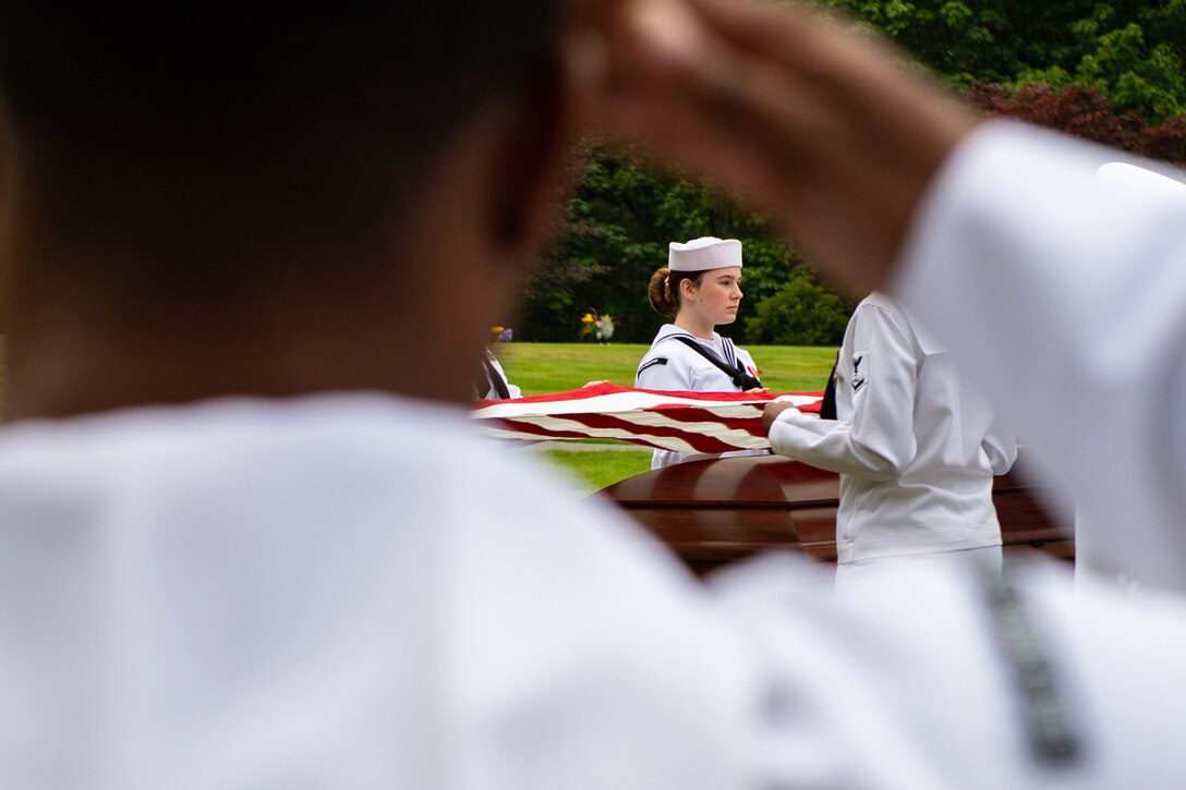 A sailor salutes as a flag is placed over a coffin during a ceremony.