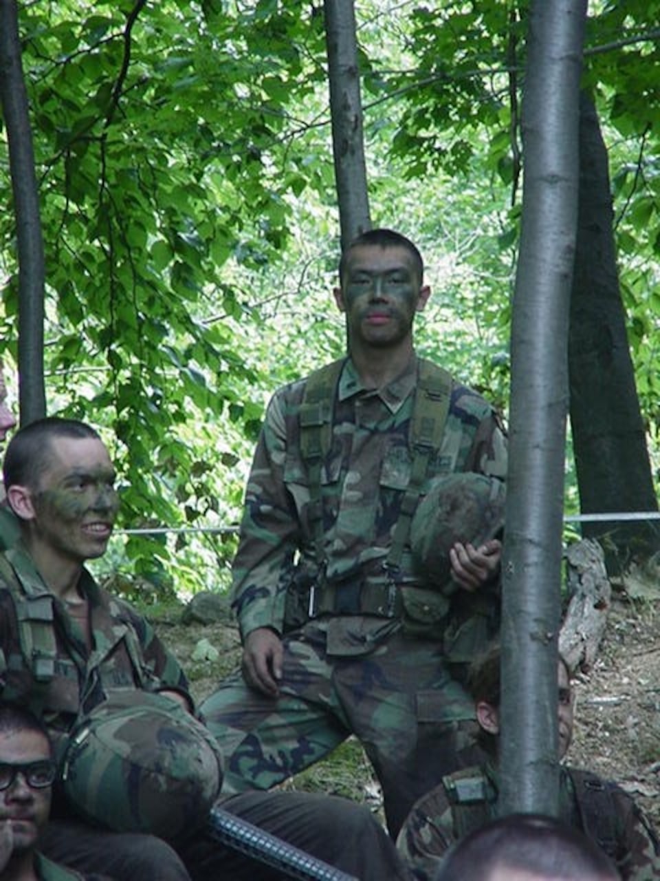 Army cadet stands alongside his fellow cadets during a field training exercise in a forested area.