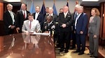 Two people seated at the end of a long conference table, looking down while signing a document with a crowd standing behind them