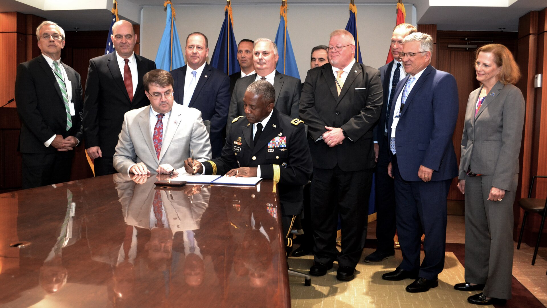 Two people seated at the end of a long conference table, looking down while signing a document with a crowd standing behind them