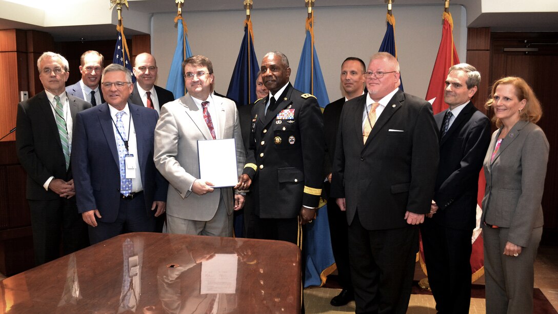 A crowd is assembled at the end of a conference table and in front of several flags, with the middle two people displaying a signed document