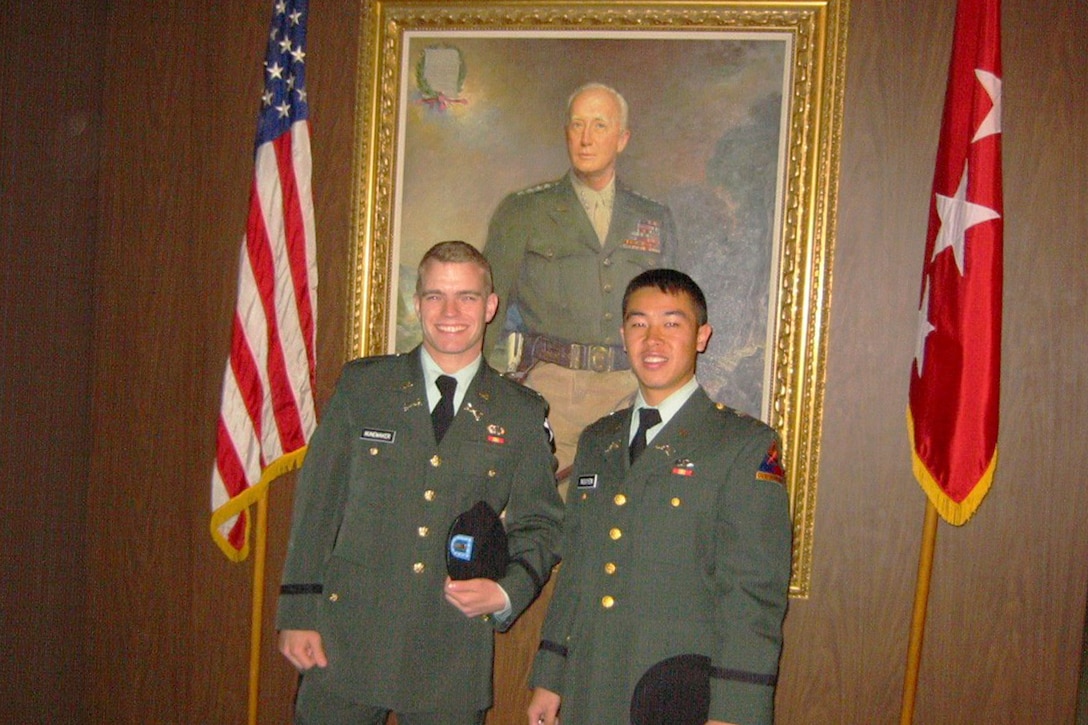 Two Army cadets pose for a photo in front of historic West Point art in their green service uniforms.