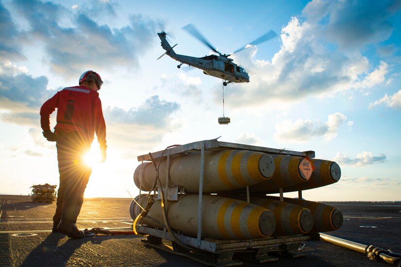 A sailor stands next to  equipment while a helicopter flies above.