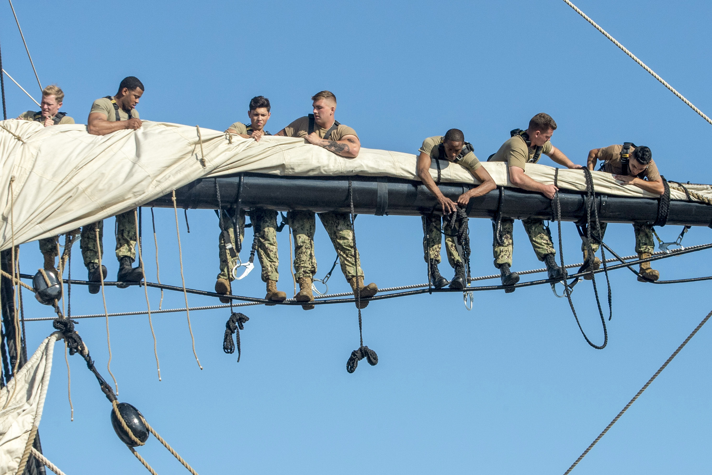 Sailors balance on the rigging while folding up a sail over the mast of a ship.