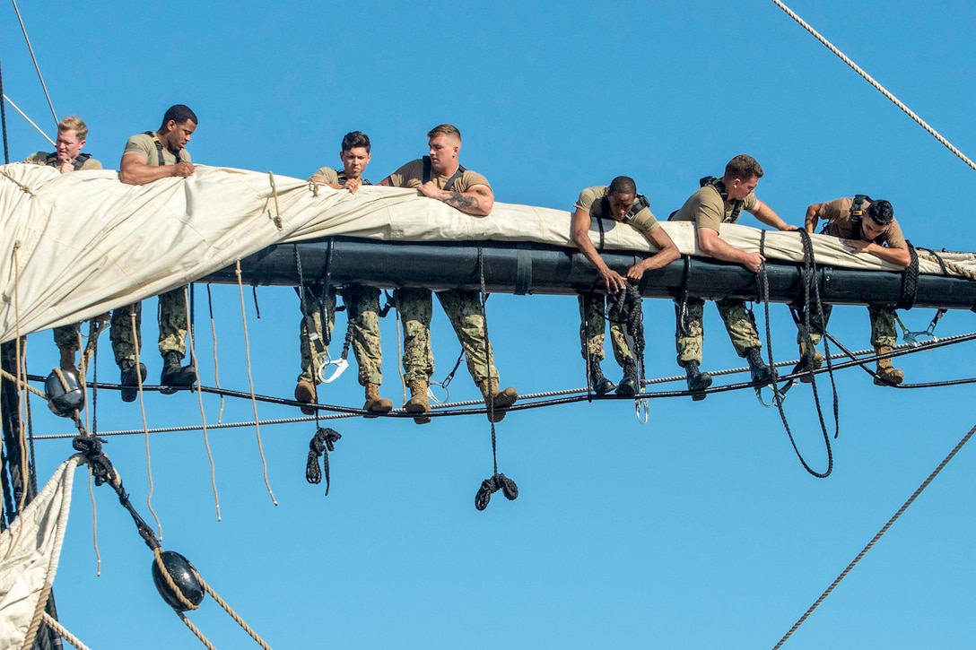 Sailors perched on a horizontal bar on a sailing vessel secure a canvas sail on it.