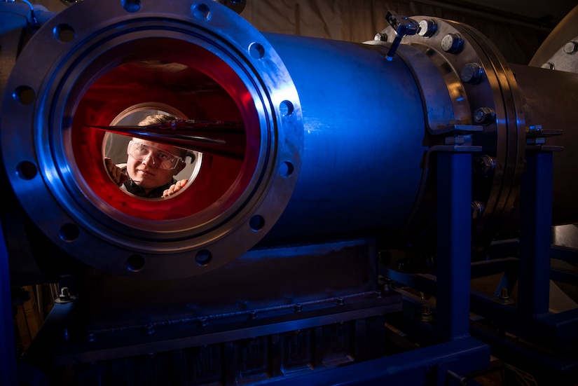 The face of a man, wearing protective glasses, is seen through windows in a large metal tube. Inside the tube is a pointed metallic object.