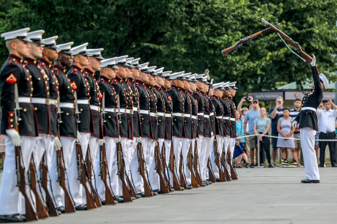 Marines stand in a line as one Marine facing them catches a rifle mid-air.