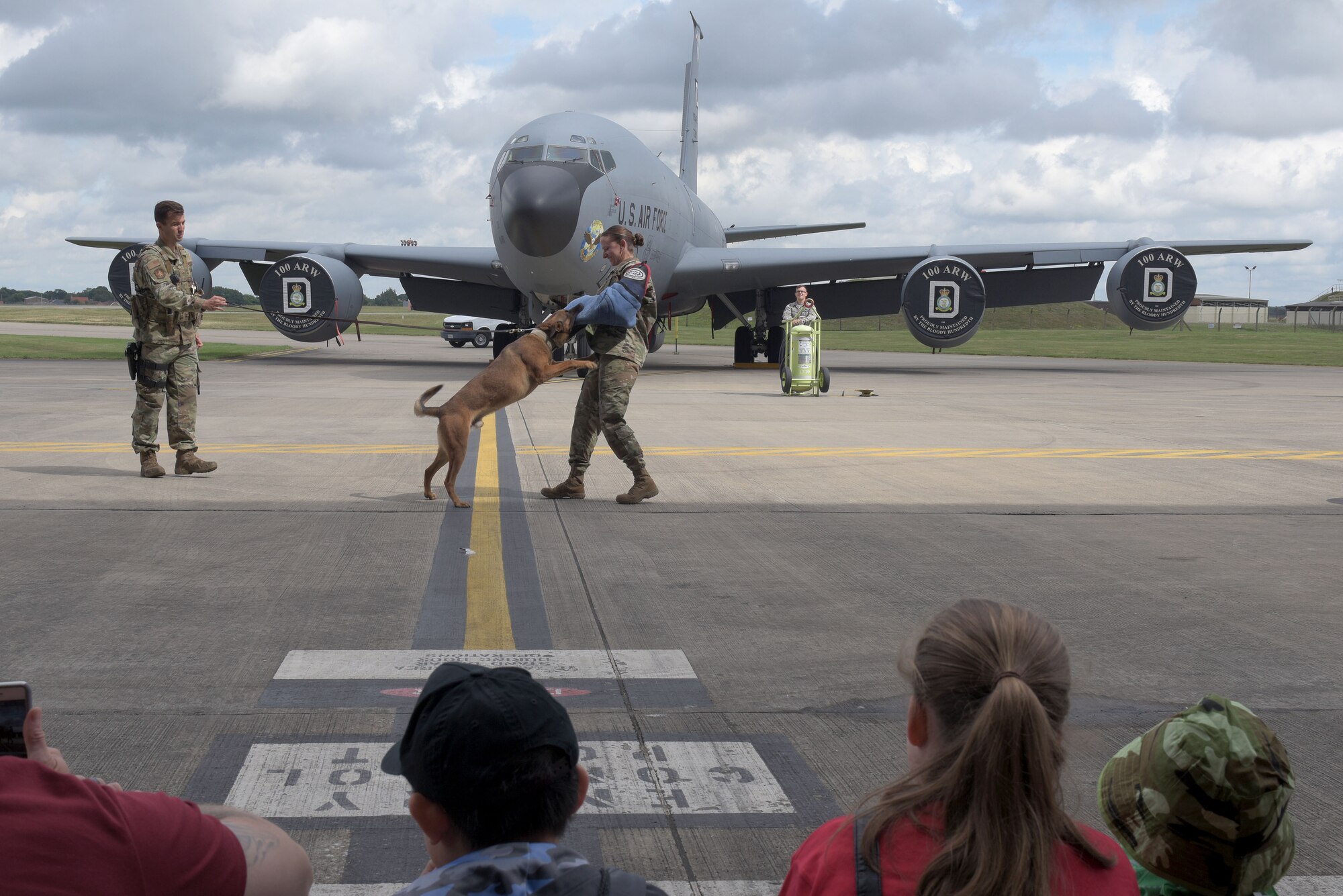 From the left: Staff Sgt. Camron Quaranto, 100th Security Forces Squadron military working dog handler, and his partner Karo perform a bite demonstration with Tech. Sgt. Melissa Burns, 100th SFS MWD kennel master, during the ‘kids deployment line’ at RAF Mildenhall, England, Aug. 9, 2019. After the demonstration the children toured a KC-135 Stratanker, a CV-22 Osprey and an MC-130J Commando II. (U.S. Air Force photo by Senior Airman Benjamin Cooper)