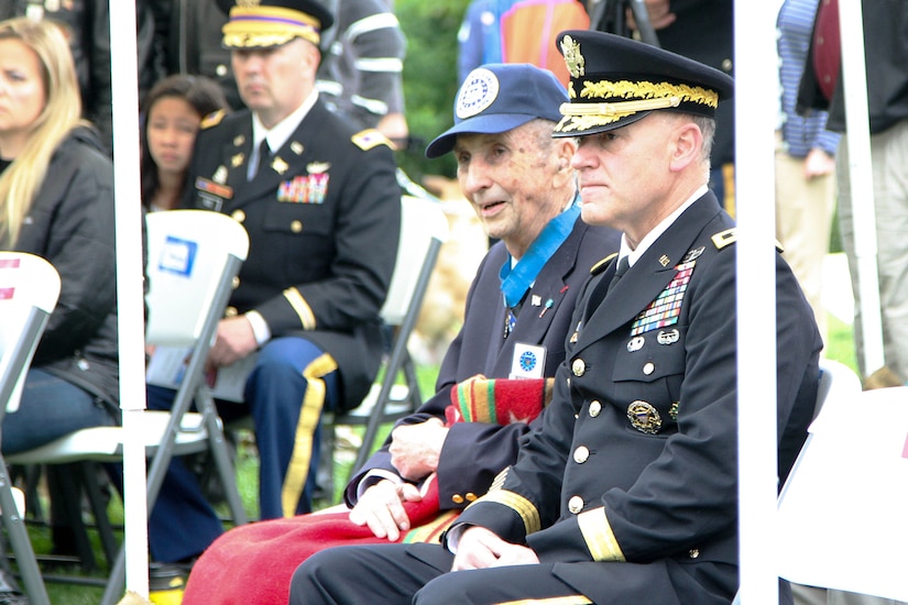 An Army lieutenant general in dress uniform sits in a folding chair next to an older veteran wearing a Medal of Honor around his neck. The older man is holding a blanket over his legs. Other people can be seen sitting in similar chairs in the background.