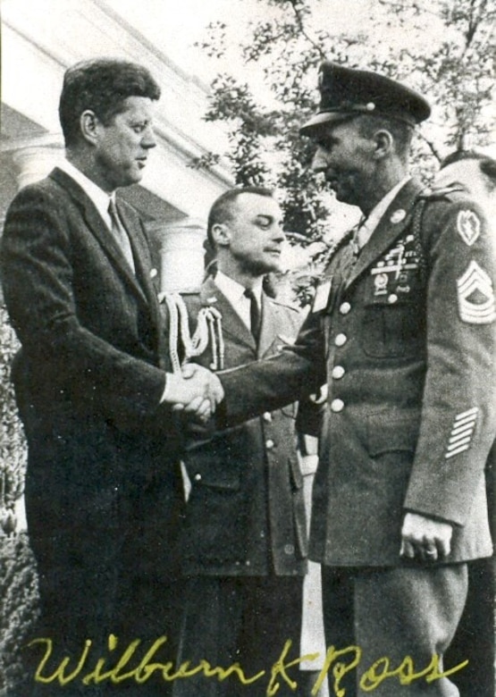 President John F. Kennedy shakes hands with an Army soldier in full dress uniform. Another soldier stands behind them looking in another direction.