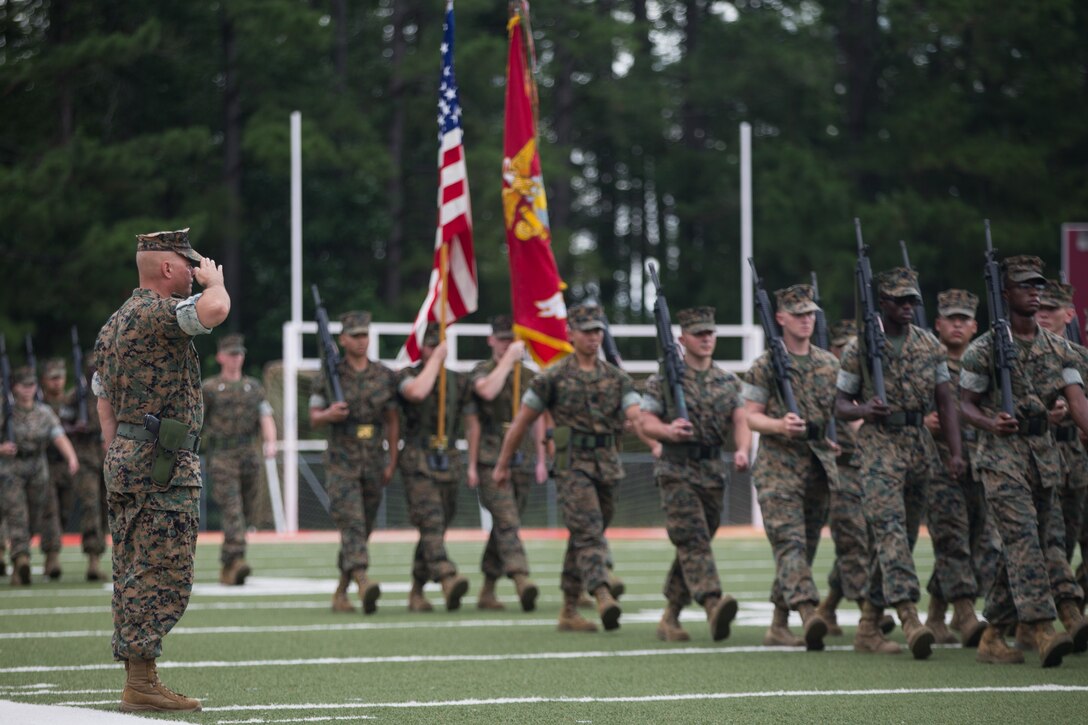 A Marine in battle dress salutes other Marines marching in formation. Those Marines are holding rifles and have a color guard following them.