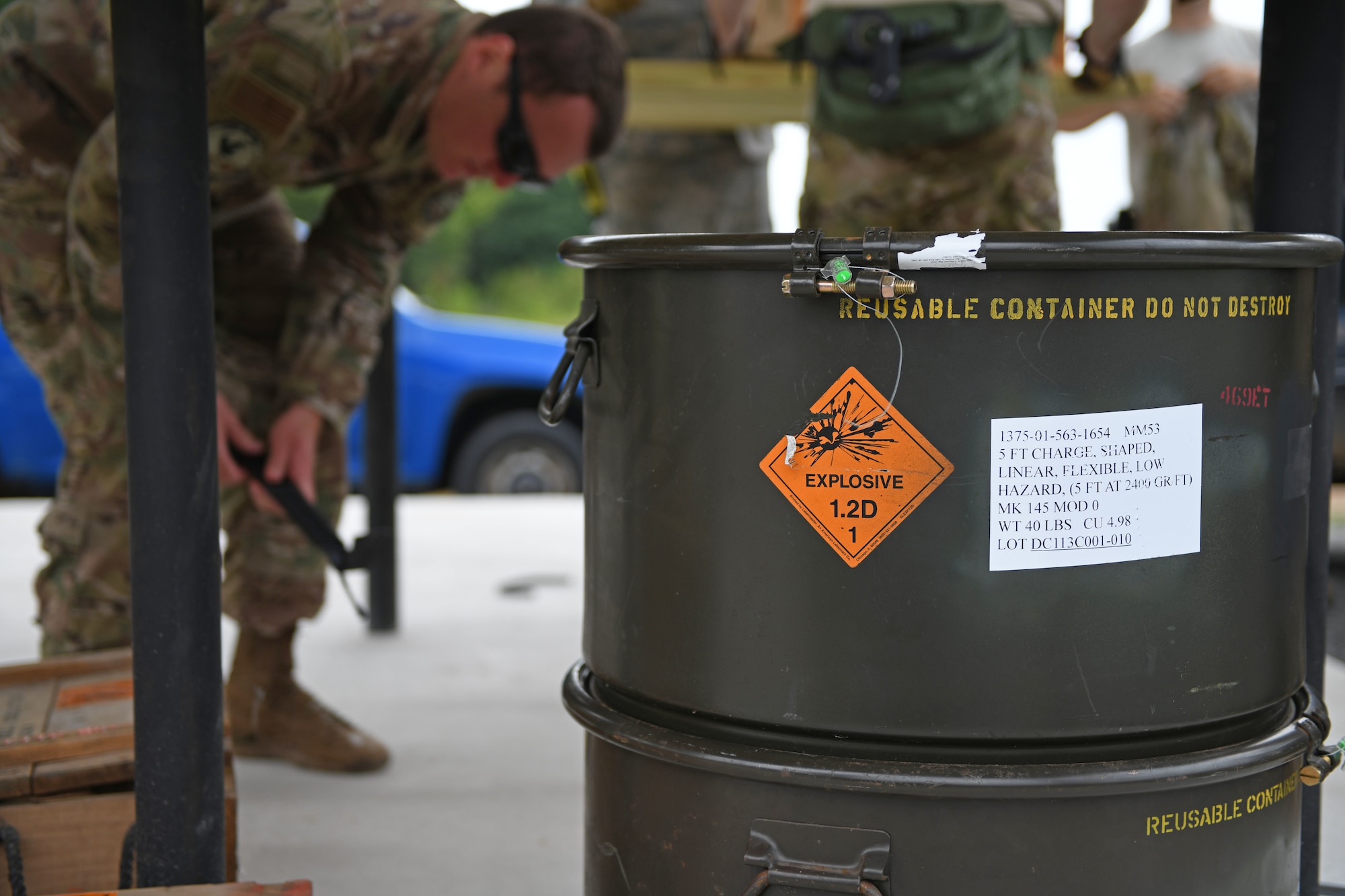 A man kneels over behing a dark green cylindrical drum which has an orange sticker on it that says explosive.