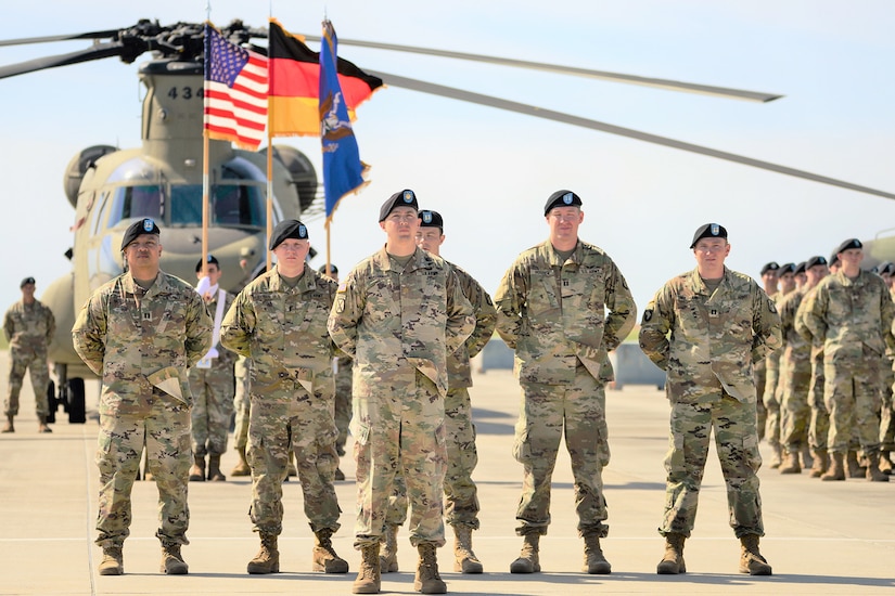 Six combat-dressed Army soldiers stand in formation with their hands behind their backs. A color guard is behind them, as is an Army helicopter. More soldiers stand in formation in the background.