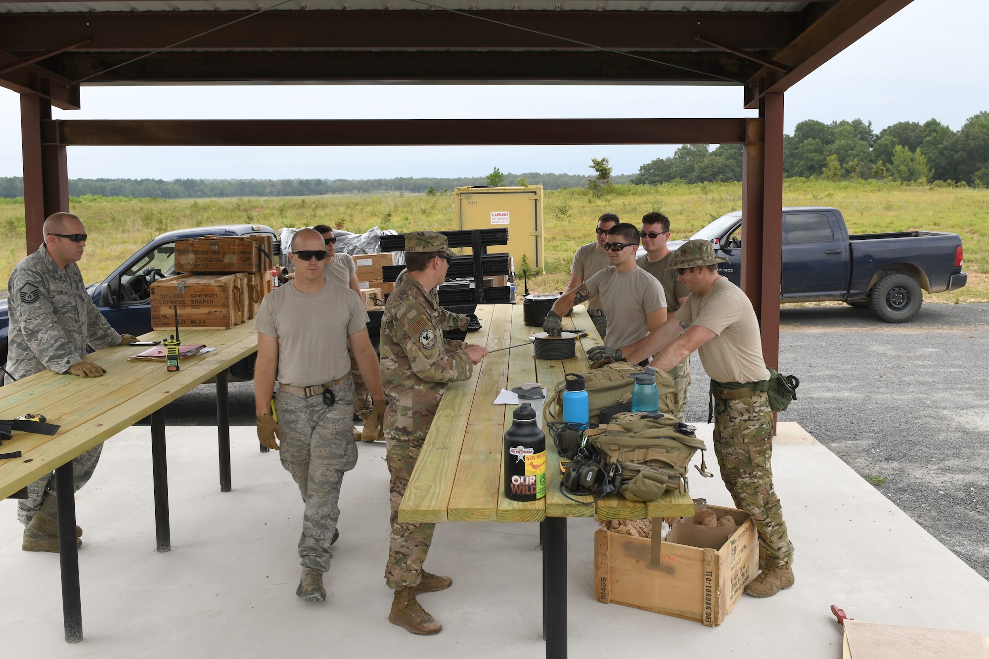 8 men stand by wooden tables with 2 blue trucks in the background.