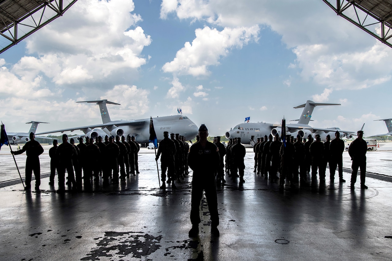 Several uniformed men whose silhouettes are shaded stand in three formations underneath an aircraft hangar. One man stands in front of them. Two large military cargo aircraft are parked behind them.