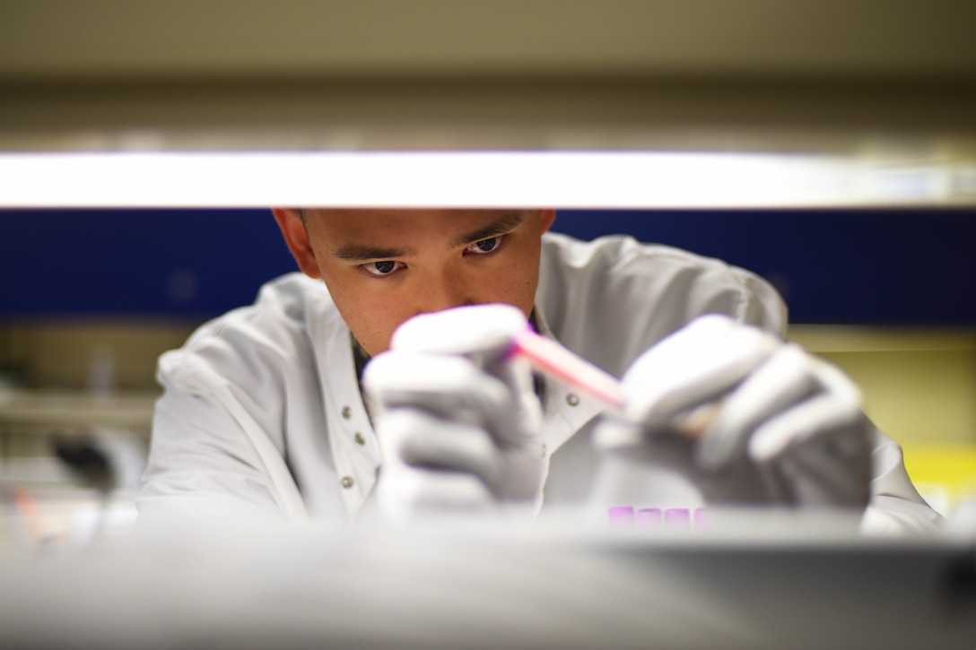 A service member in a lab coat studies a sample he's holding with white-gloved hands.
