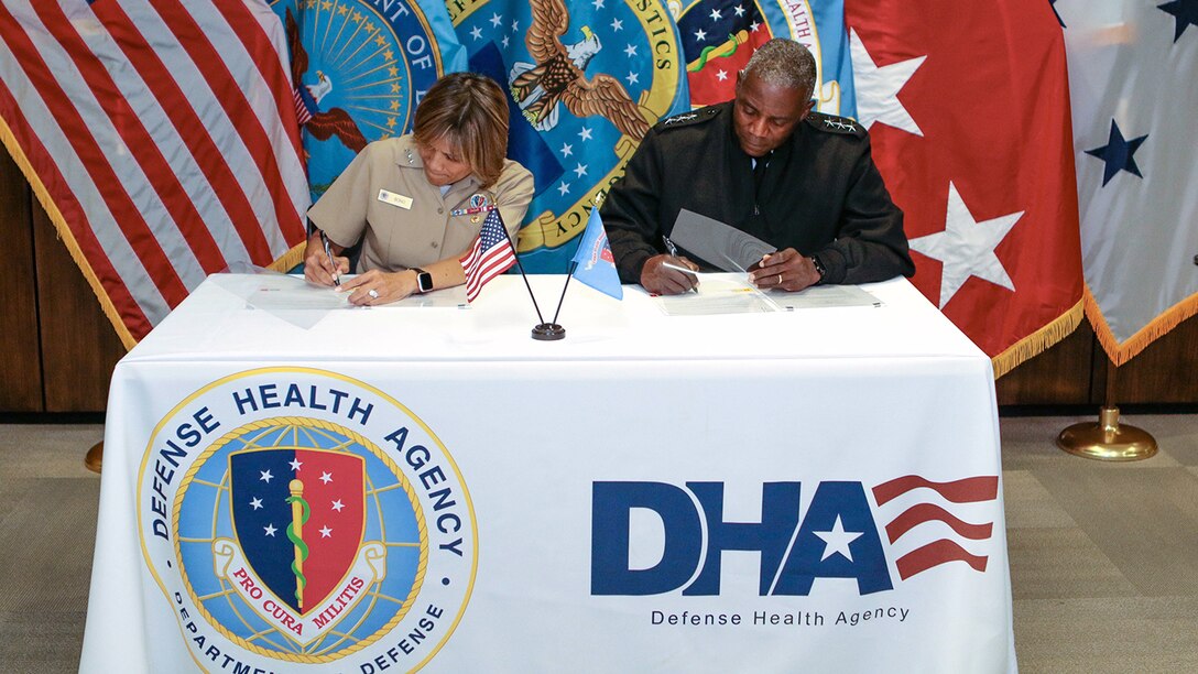 Navy Vice Adm. Raquel Bono, seated left, and Army Lt. Gen. Darrell K. Williams,seated right, sign a memorandum of agreement at a table featuring the DHA emblem
