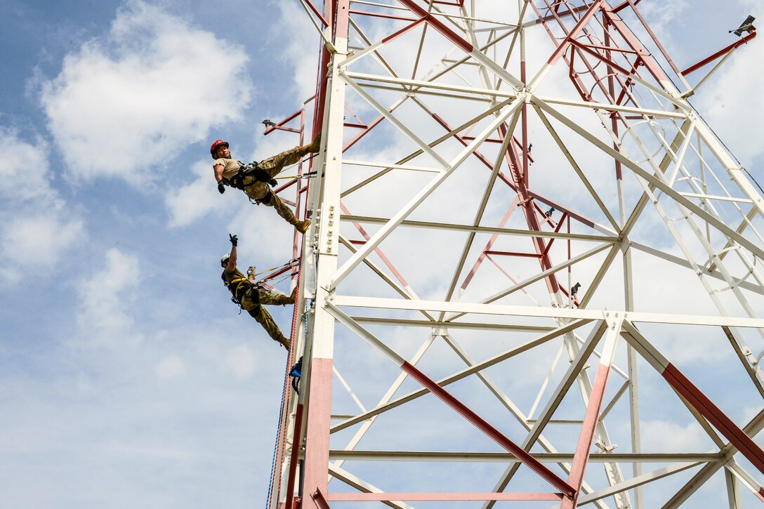 Two airmen climb a metal tower, with blue sky and white clouds in the background.