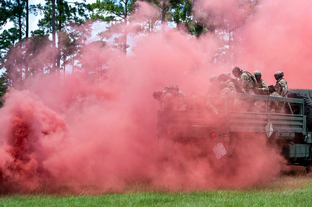 Service members in a vehicle drive on a field through a reddish-pink smoke cloud.