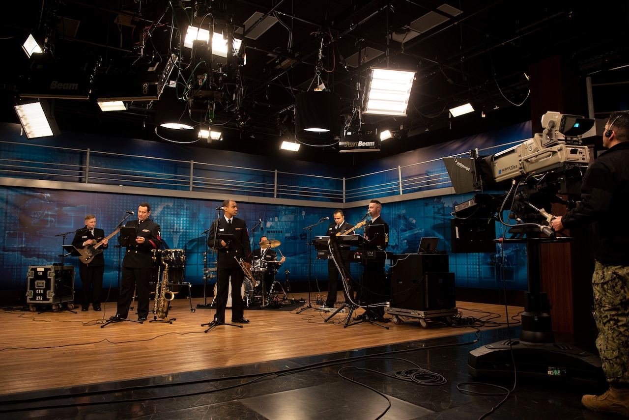 Musicians play instruments in a studio on a stage with overhead lights.