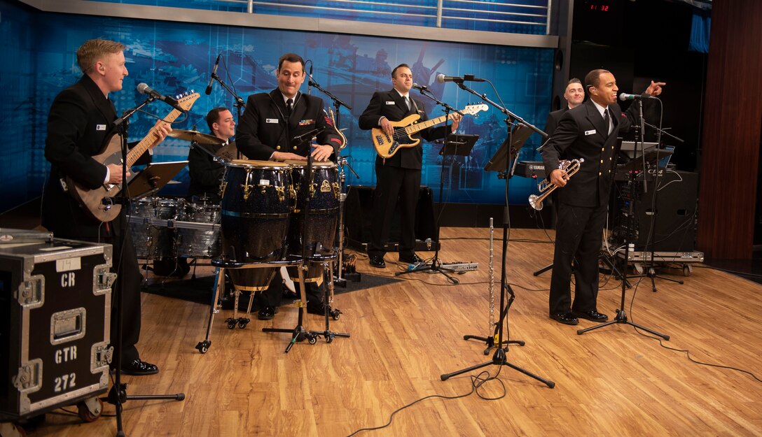 Men in Navy uniforms play instruments and sing into microphones.