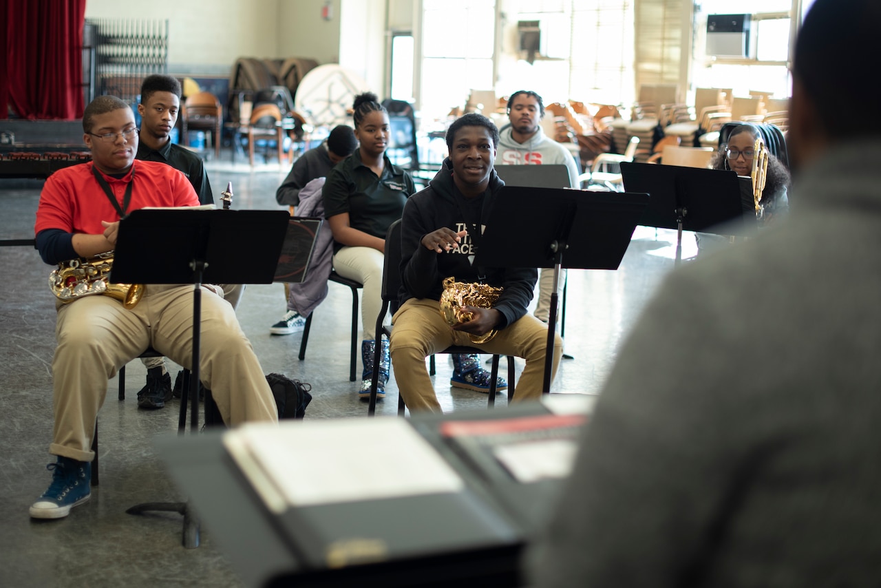 Students holding instruments with music stands in front them sit in a classroom.