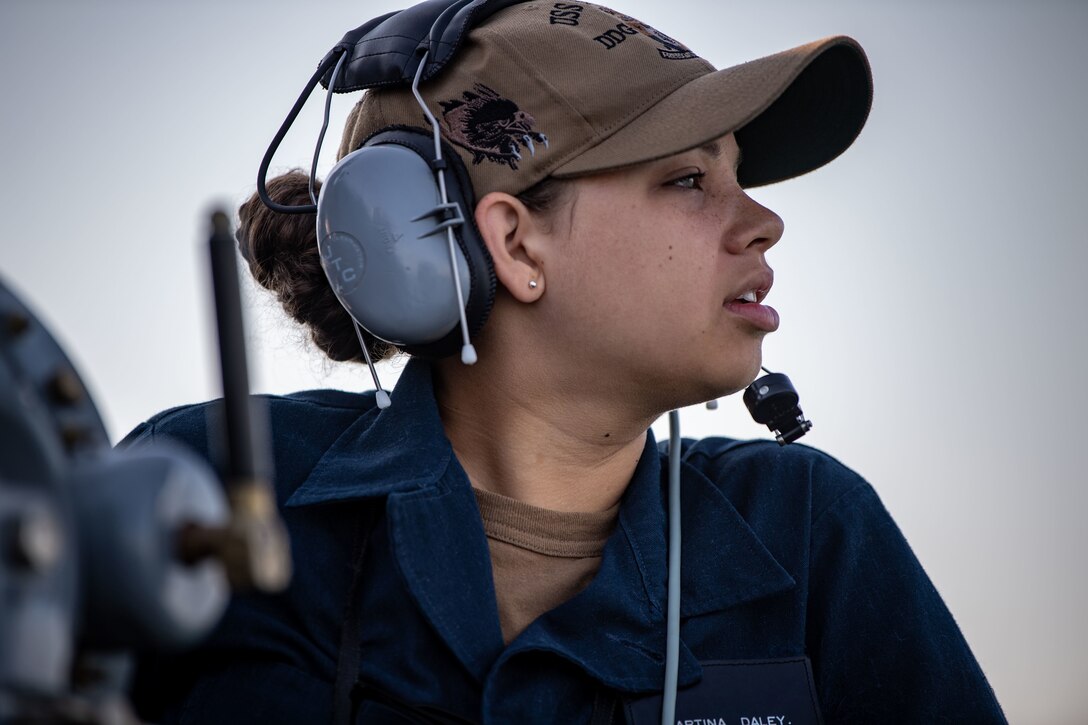 A female sailor with a headset on looks to the right.