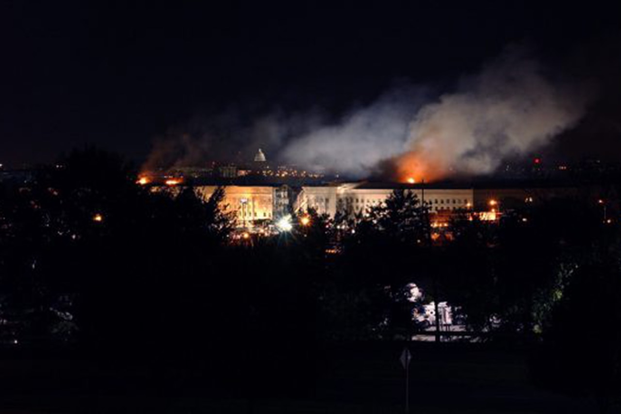 A night time shot of a  building in the distance with  smoke and flames coming from it.