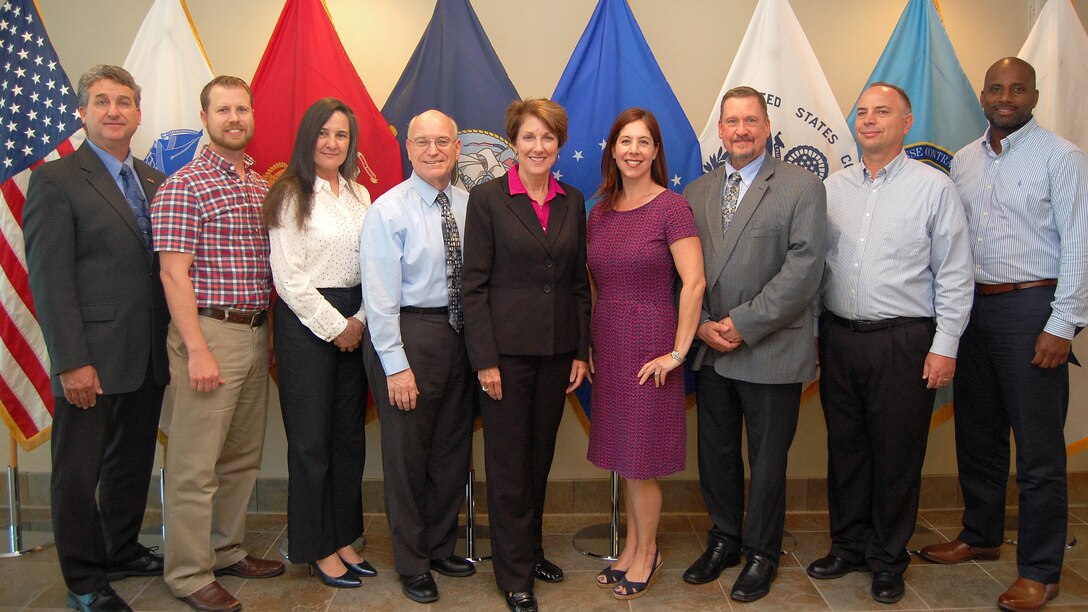 A group of DCMA employees stand in front of flags.