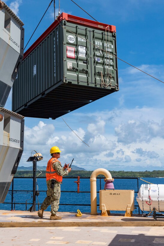 A man in military uniform wears a hard hat and carries a portable radio.  Above him is a cargo container suspended from a crane.