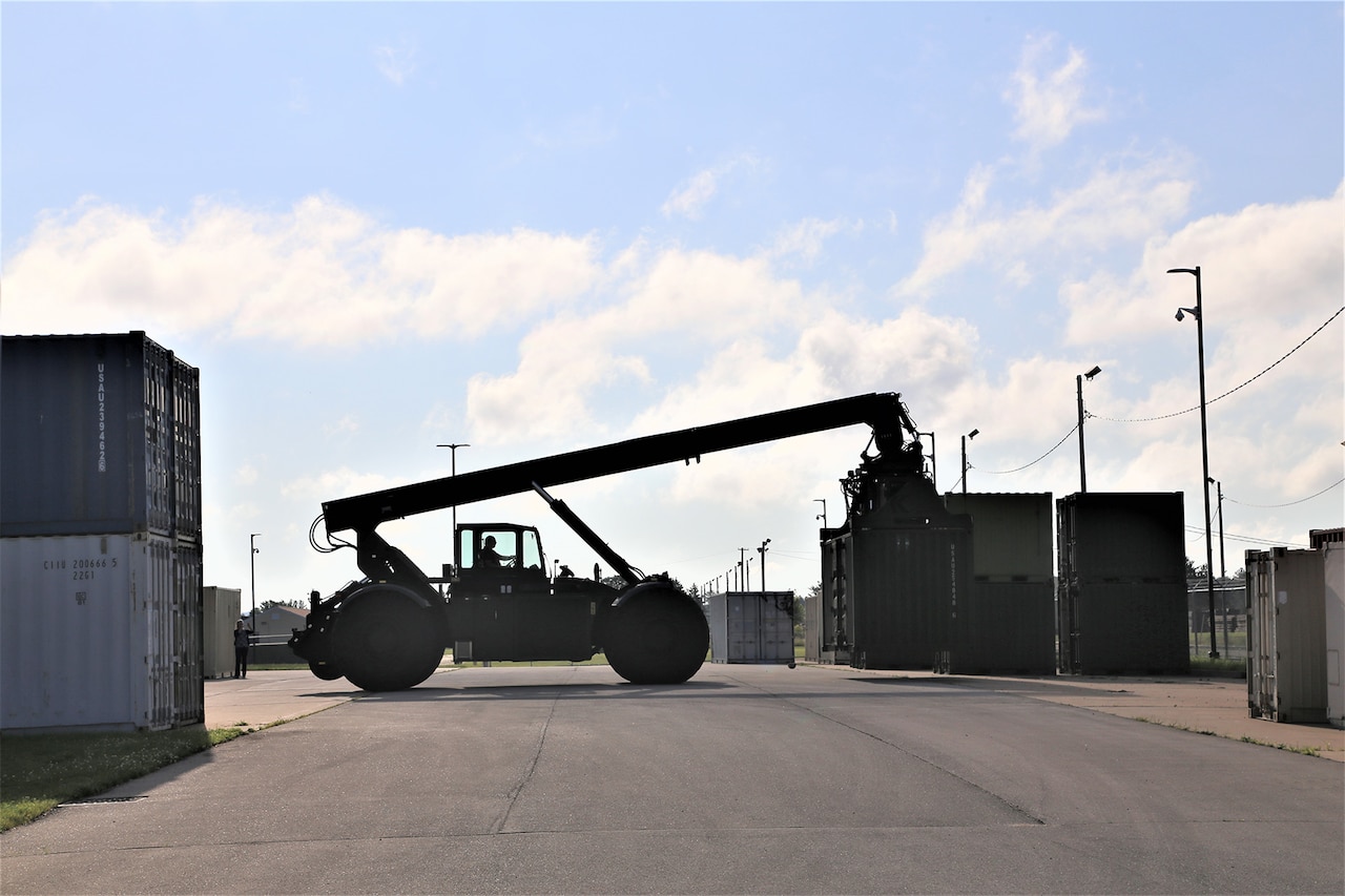 A large industrial vehicle in silhouette against a bright blue sky picks up a shipping container.