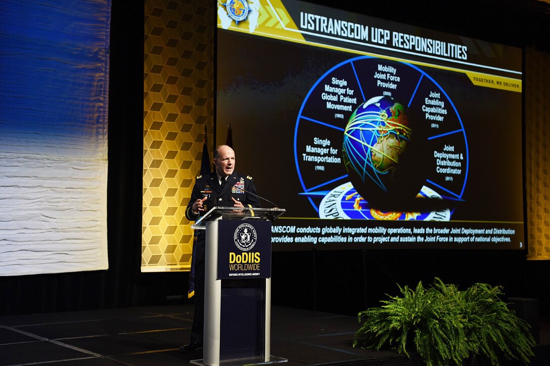 A man in military uniform stands behind a lectern. On the wall behind him is a projection that reads “USTRANSCOM UCP Responsibilities.”