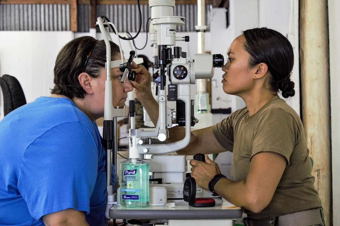 An optometrist sits on one side of an optical device and examines a patient sitting across from her.