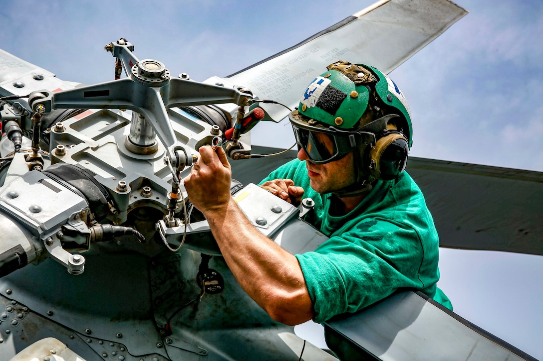 A sailor wearing green manipulates a helicopter rotor.