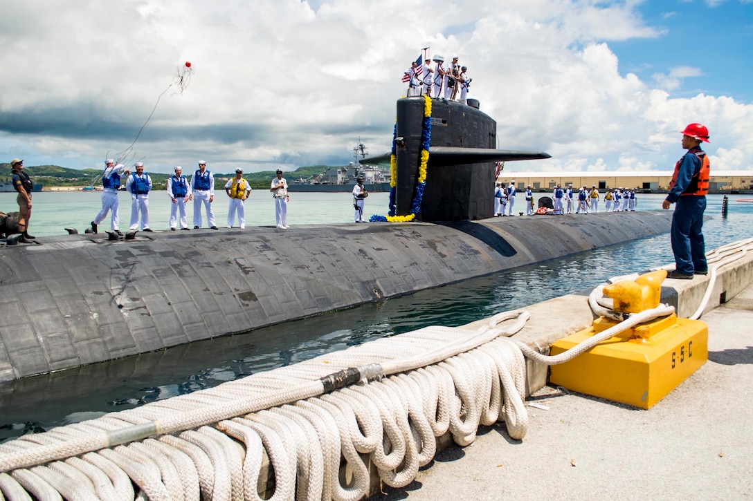 Sailors stand on top of a submarine as it is near a pier.