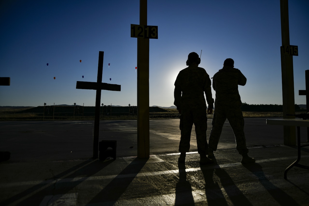 Two airmen silhouetted as they stand near some cross-shaped posts, with airborne drones in the sky.