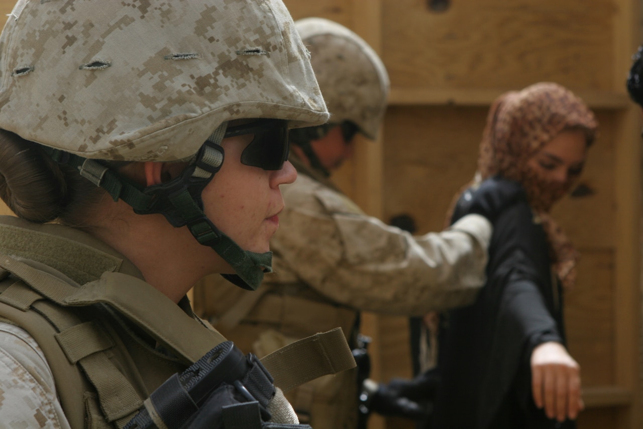 A Marine in combat equipment stands in the foreground as another Marine searches a role-playing civilian.
