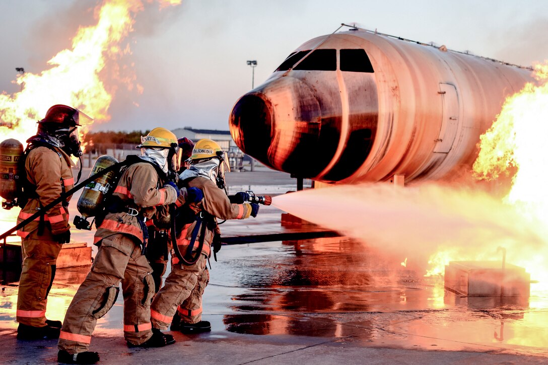 Two firefighters spray water on an aircraft fire as a third one watches.