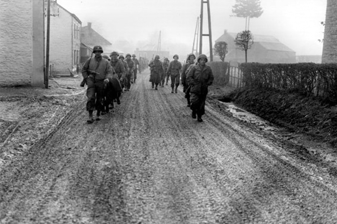 Soldiers in lines of two march down a muddy street. In the background are houses, a tree and several poles.