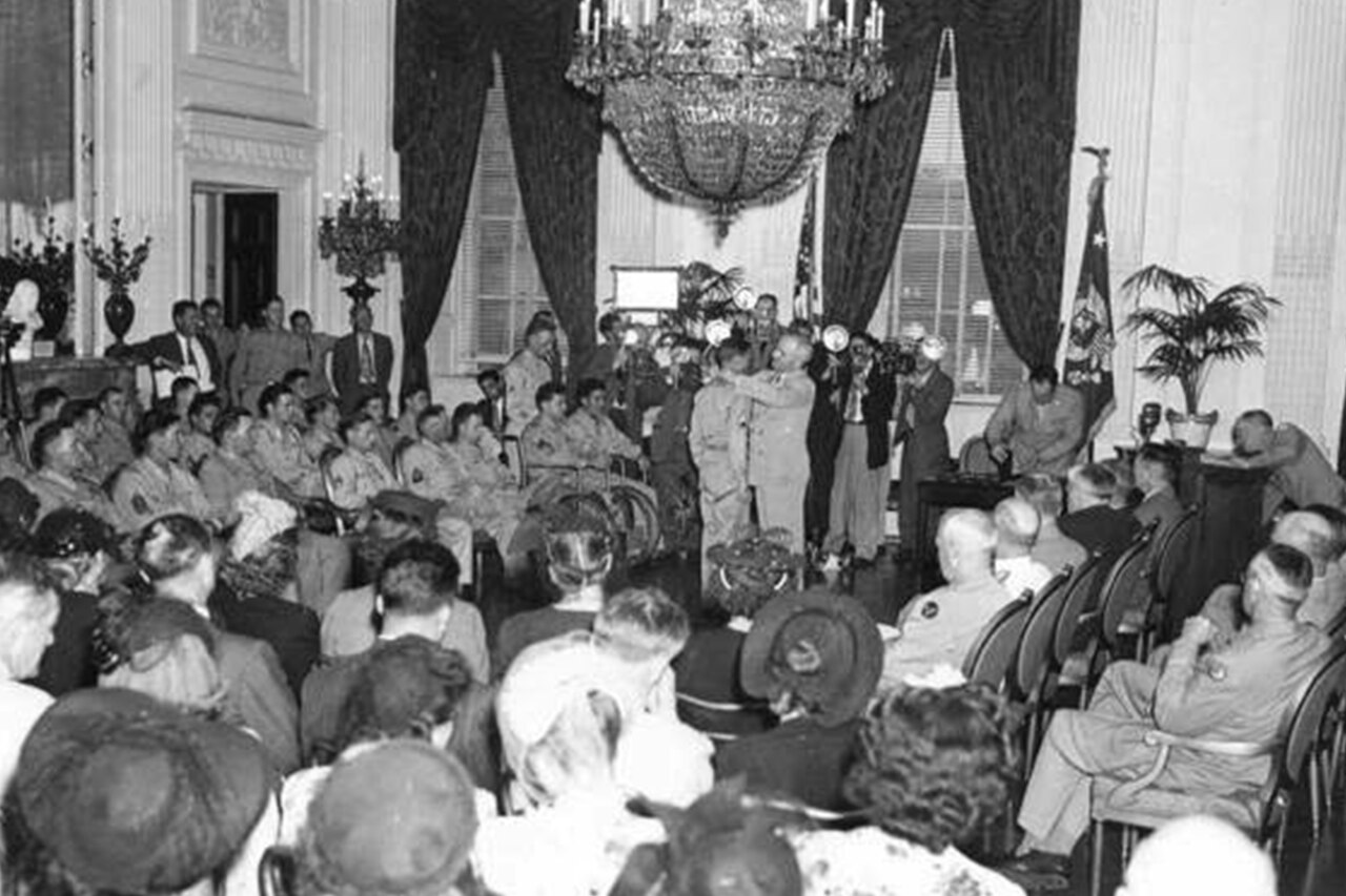 In a room with high ceilings, two curtained windows and a chandelier, a man places a medal around a soldier’s neck. Several other soldiers and civilians watch, seated.