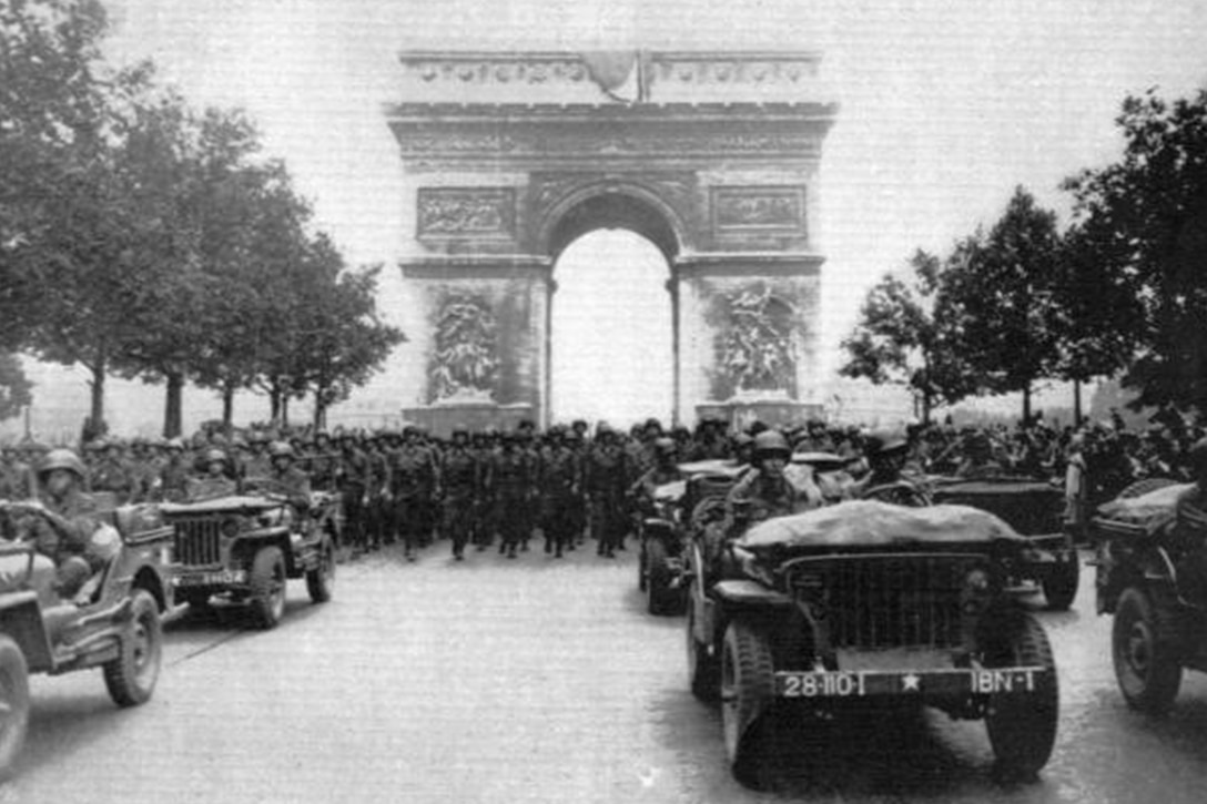 Five Army vehicles drive down a tree-lined street, with hundreds of soldiers marching behind them. In the background is the famed Parisian Arc de Triumphe.