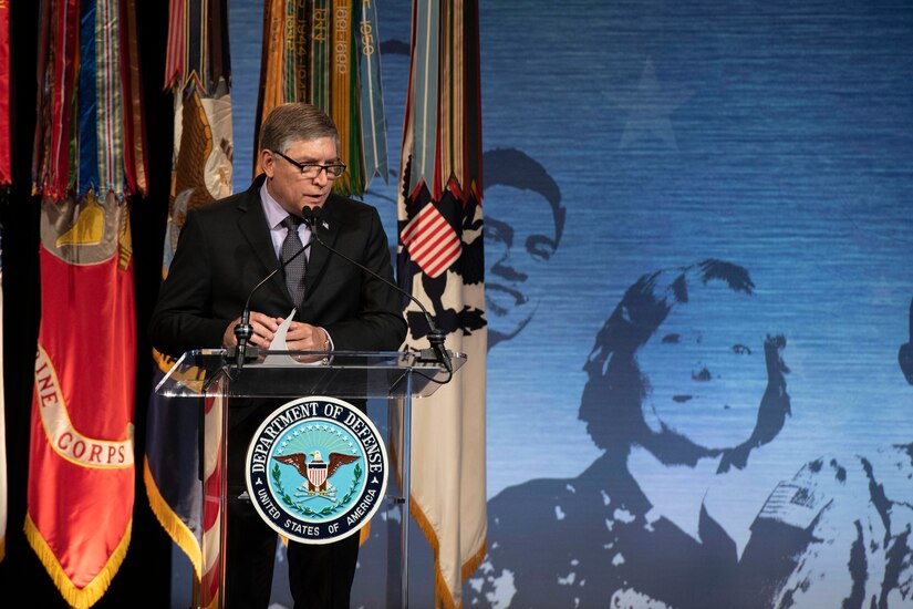 A man stands behind a lectern that bears the seal of the Department of Defense. Behind him are four military flags.