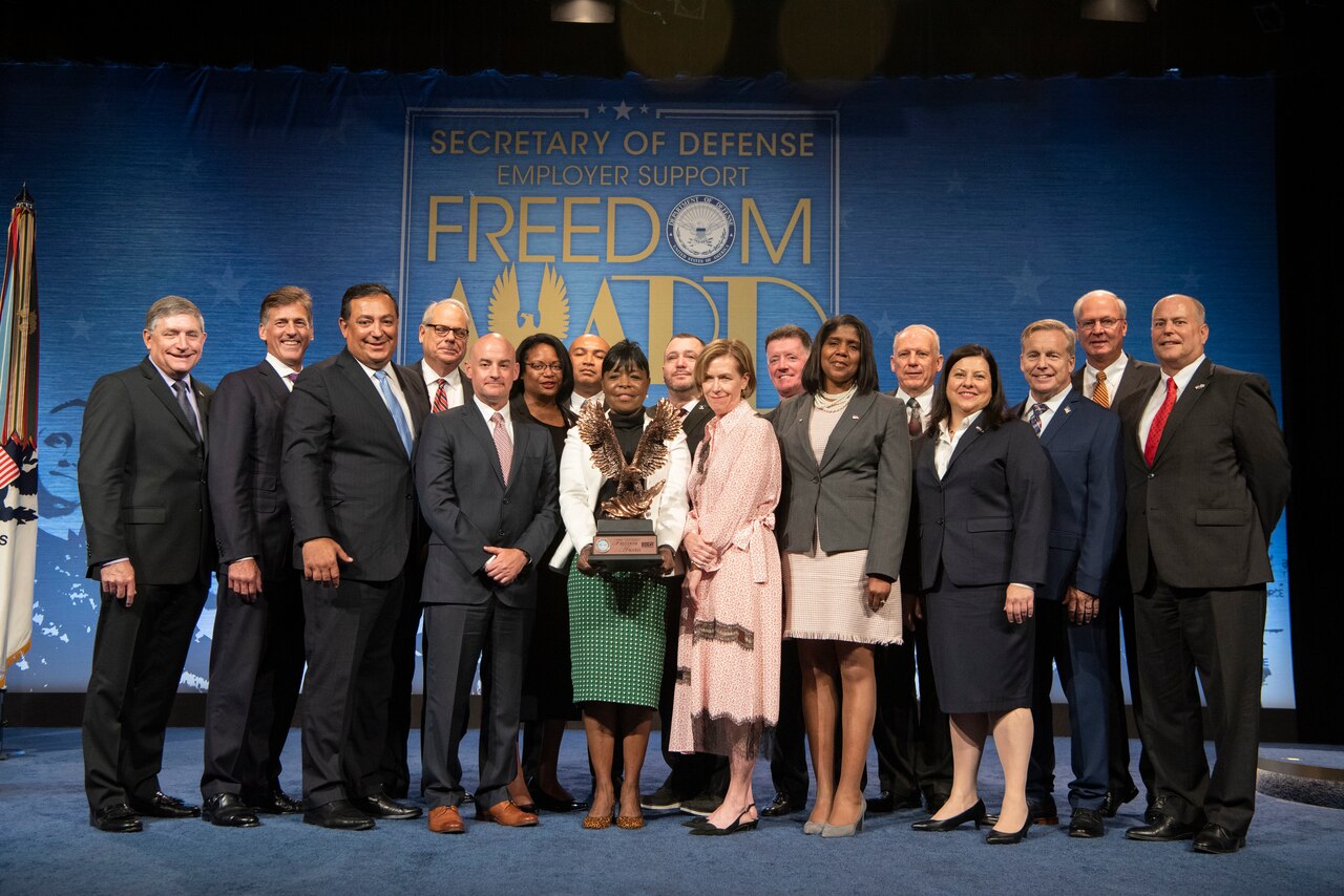 Seventeen individuals in professional attire stand together on a stage. In the center, a woman holds an eagle-shaped trophy.