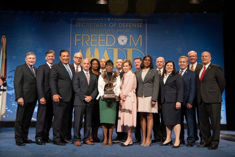 Seventeen individuals in professional attire stand together on a stage. In the center, a woman holds an eagle-shaped trophy.