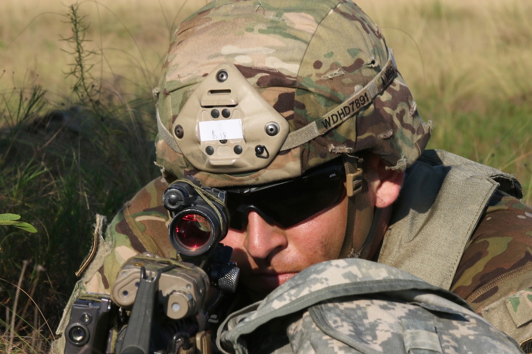 A soldier low in the grass peers through a weapon scope.