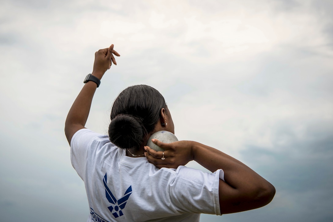 An airman, shown from behind, holds a shot put in one hand and holds the other arm up.