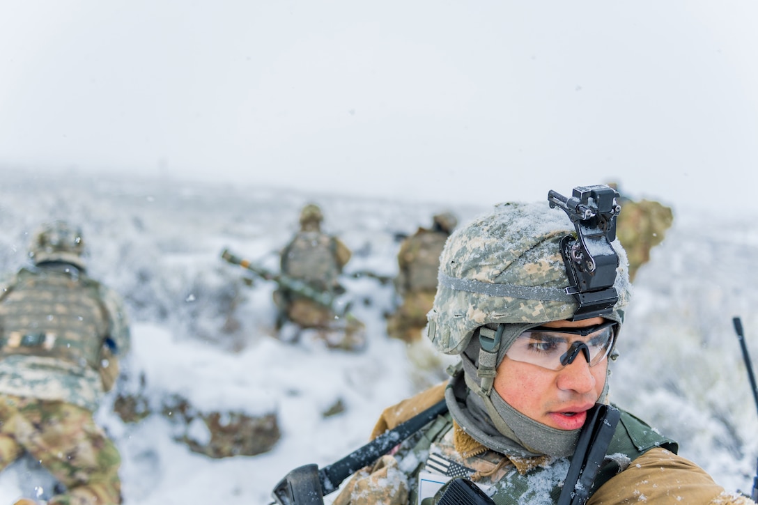 A soldier in a snow field observes something while others kneel nearby.