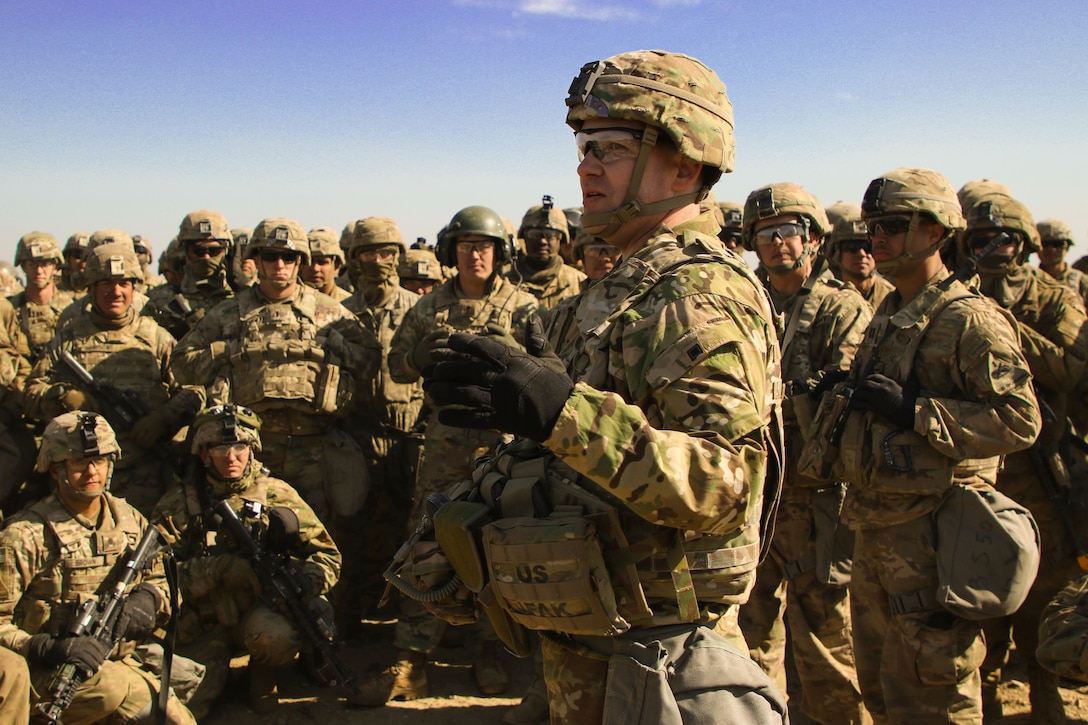 A soldier addresses a group of soldiers at an outdoor training area.