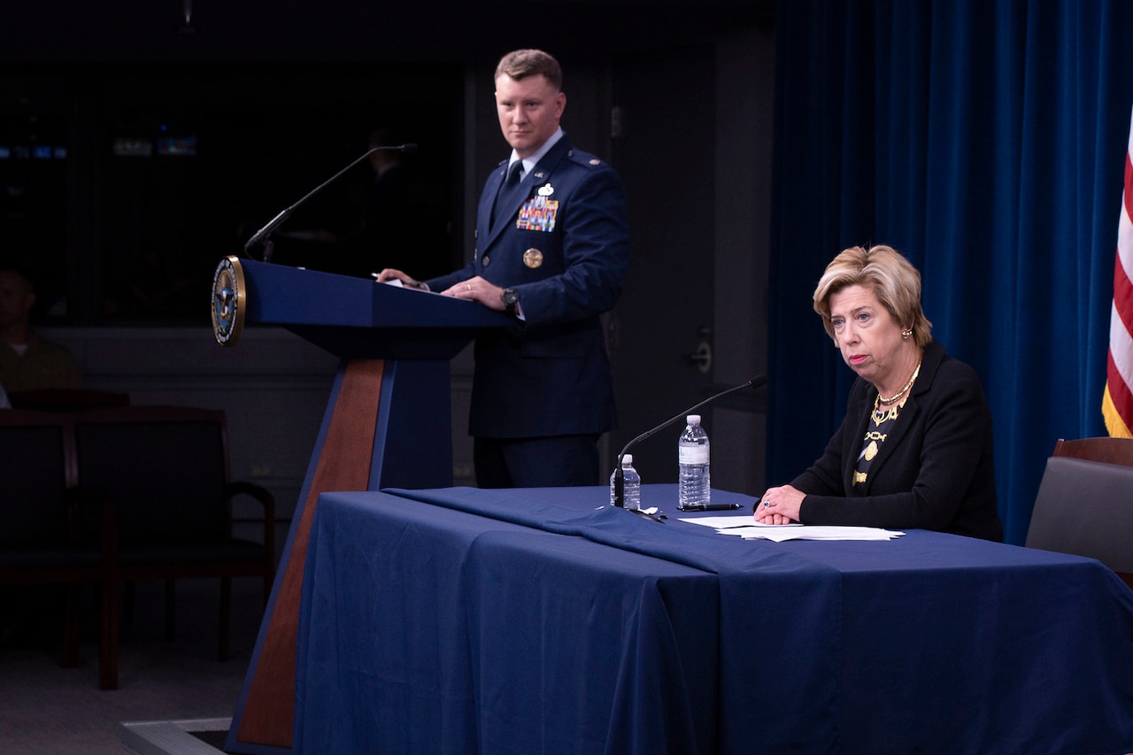 A woman sits behind a desk on a podium and speaks to reporters as a man in a military uniform stands next to her.